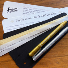 Load image into Gallery viewer, Bookmark making set resources with blank black bookmarks and gold and silver pens on a wooden background.
