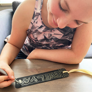 Girl using silver marker pen to design a bookmark with gold ribbon.
