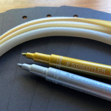 Load image into Gallery viewer, Blank black bookmarks, gold and silver pens and gold and silver ribbons on a wooden background.