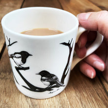 Load image into Gallery viewer, White fine bone china mug with two magpies design being held on a wooden background