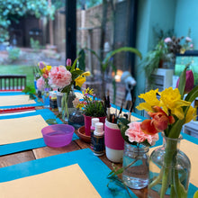 Load image into Gallery viewer, Table setup with flowers, paint supplies, and a garden view
