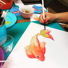 Load image into Gallery viewer, Person painting a colorful fish on paper with art supplies around
