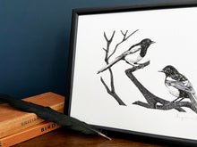 Load image into Gallery viewer, Two for joy magpies giclée print in a black frame, standing on a wooden surface with navy blue wall behind. A black feather and two bird books sit next to the print.

