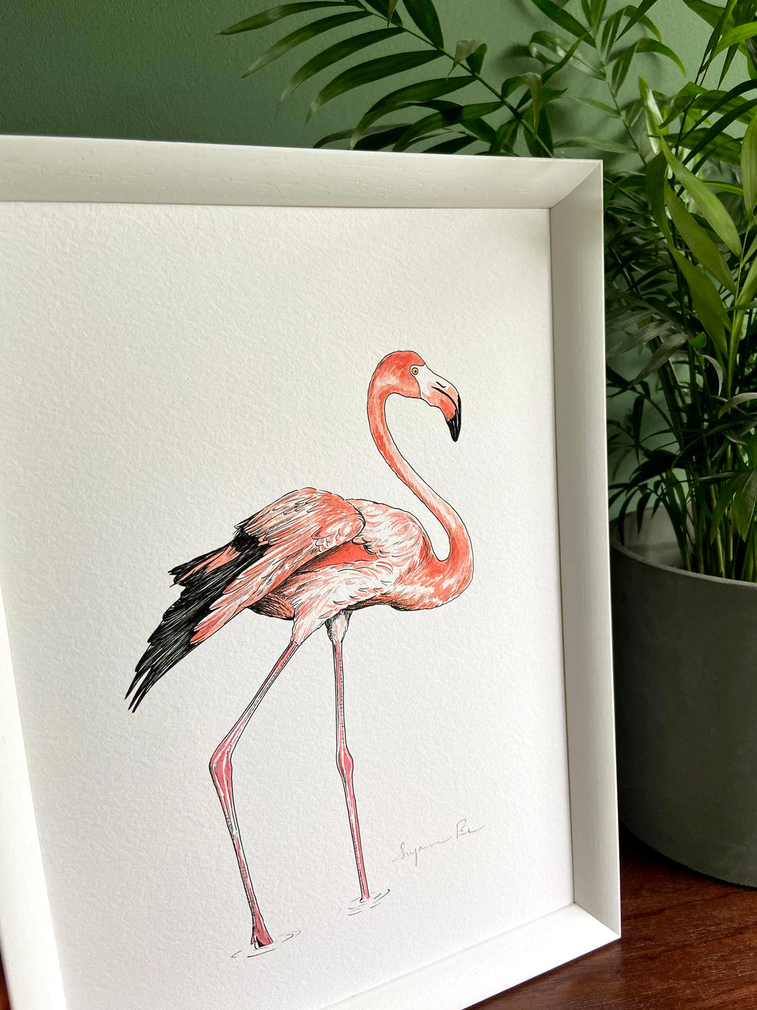 Flamingo giclée print with handpainted watercolour plumage, in a white frame, standing on a wooden surface. Green wall behind. Pot plant standing next to it.