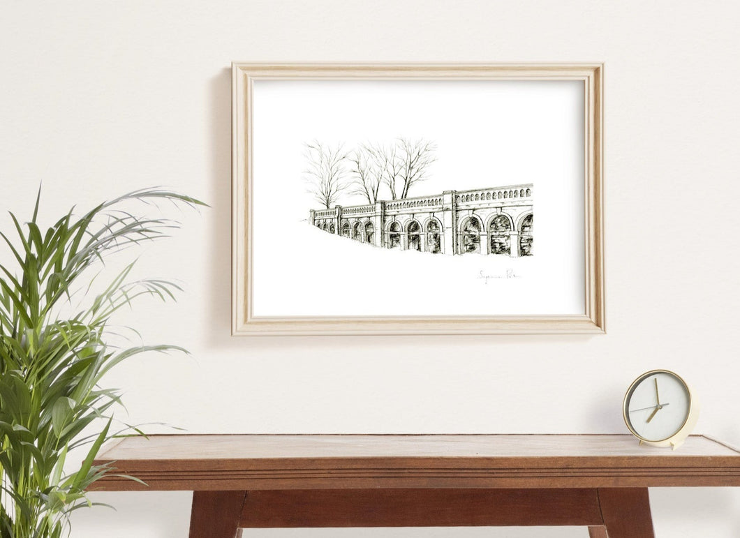 Detailed monochrome fineliner giclée print of the arches of the Italian Terraces, Crystal Palace Park, framed on a wall with wooden table, plant and clock.