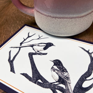 Art workshop gift card with magpies design next to a pink mug on a wooden table
