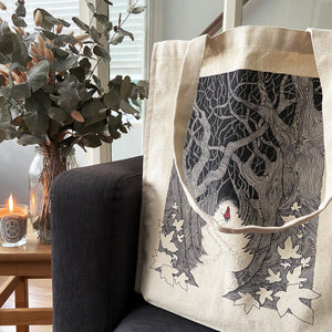 Red Riding Hood illustration canvas tote bag on a dark grey sofa next to a table with eucaplyptus leaves and a Feu de Bois candle.