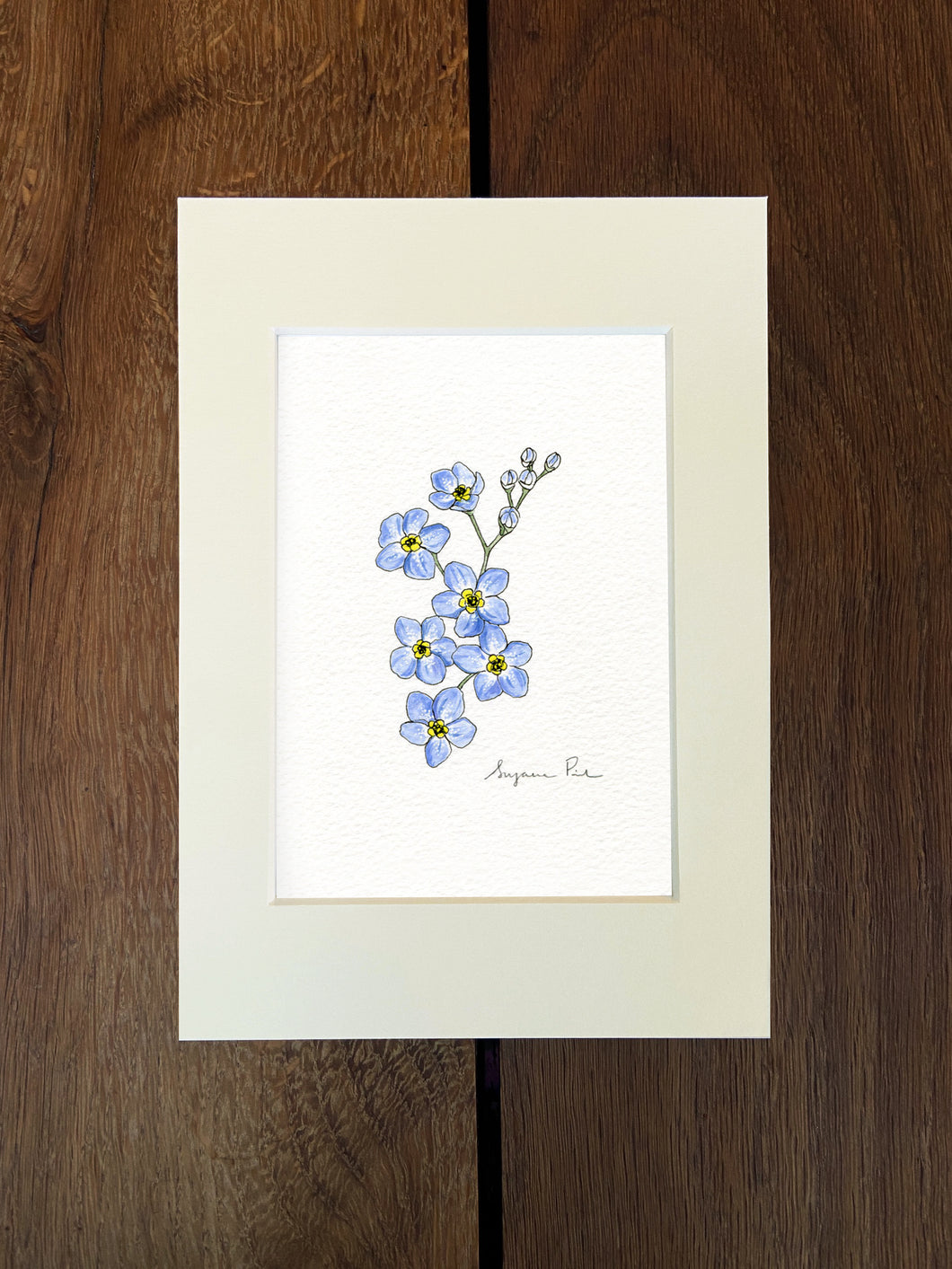 Handpainted forget-me-not design using fineliner, watercolour and white ink in an ivory mount on a wooden background.