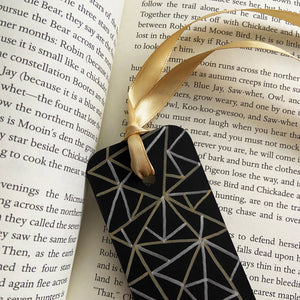 Black bookmark with gold and silver geometric pattern design and a gold ribbon on an open book.